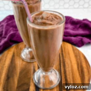Two chocolate milkshakes served in classic soda fountain glasses on a wood platter, showing off the thick, frozen texture and striped straws.