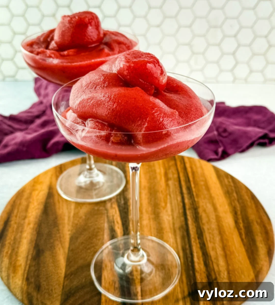 Two coupe glasses filled with thick, vibrant red frosé on a round wooden board with a purple cloth in the background.