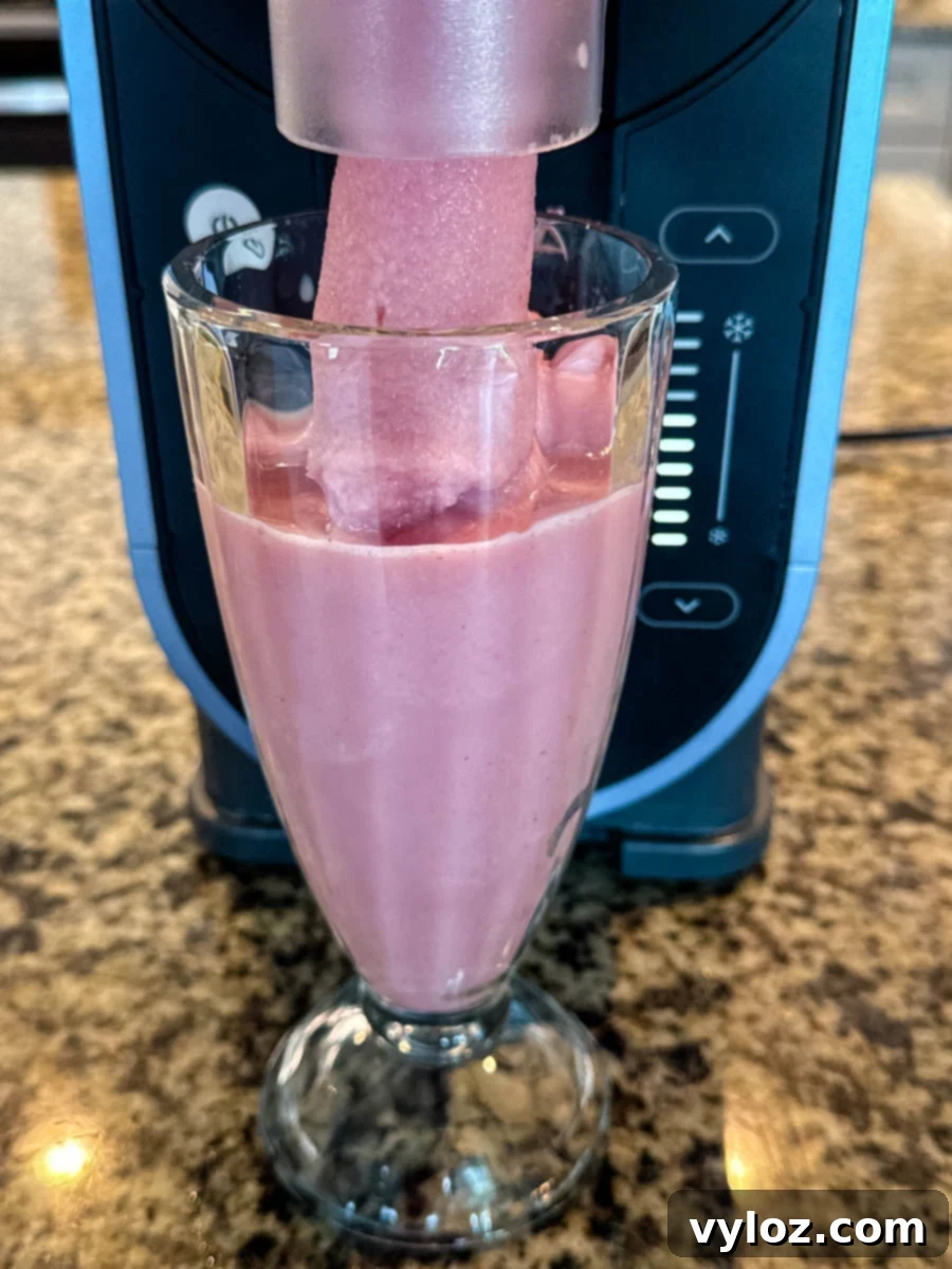 Ninja Slushi Strawberry Burst 7 A tall glass being filled with a frozen strawberry milkshake mixture directly from the Ninja Slushi machine’s spout, showing the soft, frozen texture as it pours in.