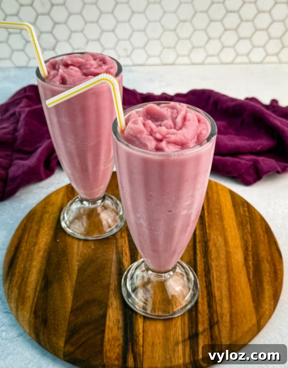 Ninja Slushi Strawberry Burst 8 Two tall glasses filled with thick, slushy pink strawberry milkshakes without whipped cream, placed on a round wooden board with a hexagonal tile background.