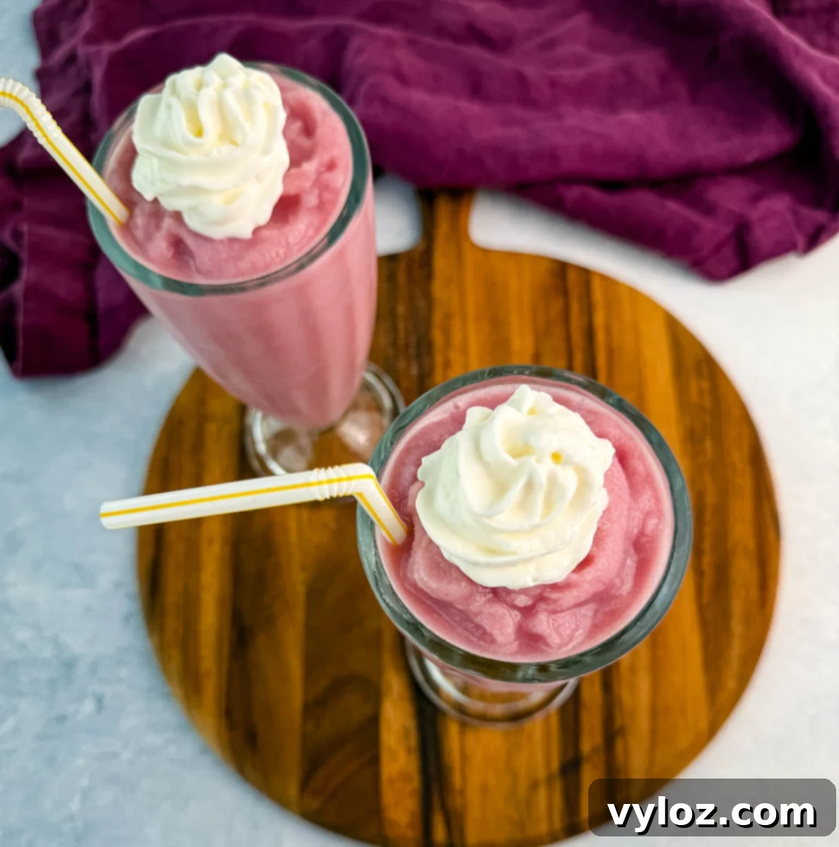 Ninja Slushi Strawberry Burst 9 Overhead view of two pink milkshakes topped with whipped cream and straws, sitting on a wooden board with a purple cloth to the side.