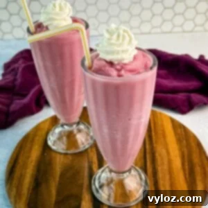 Ninja Slushi Strawberry Burst 10 Two tall milkshake glasses filled with thick pink strawberry milkshakes, topped with whipped cream and yellow-striped straws, placed on a round wooden board with a purple cloth in the background.