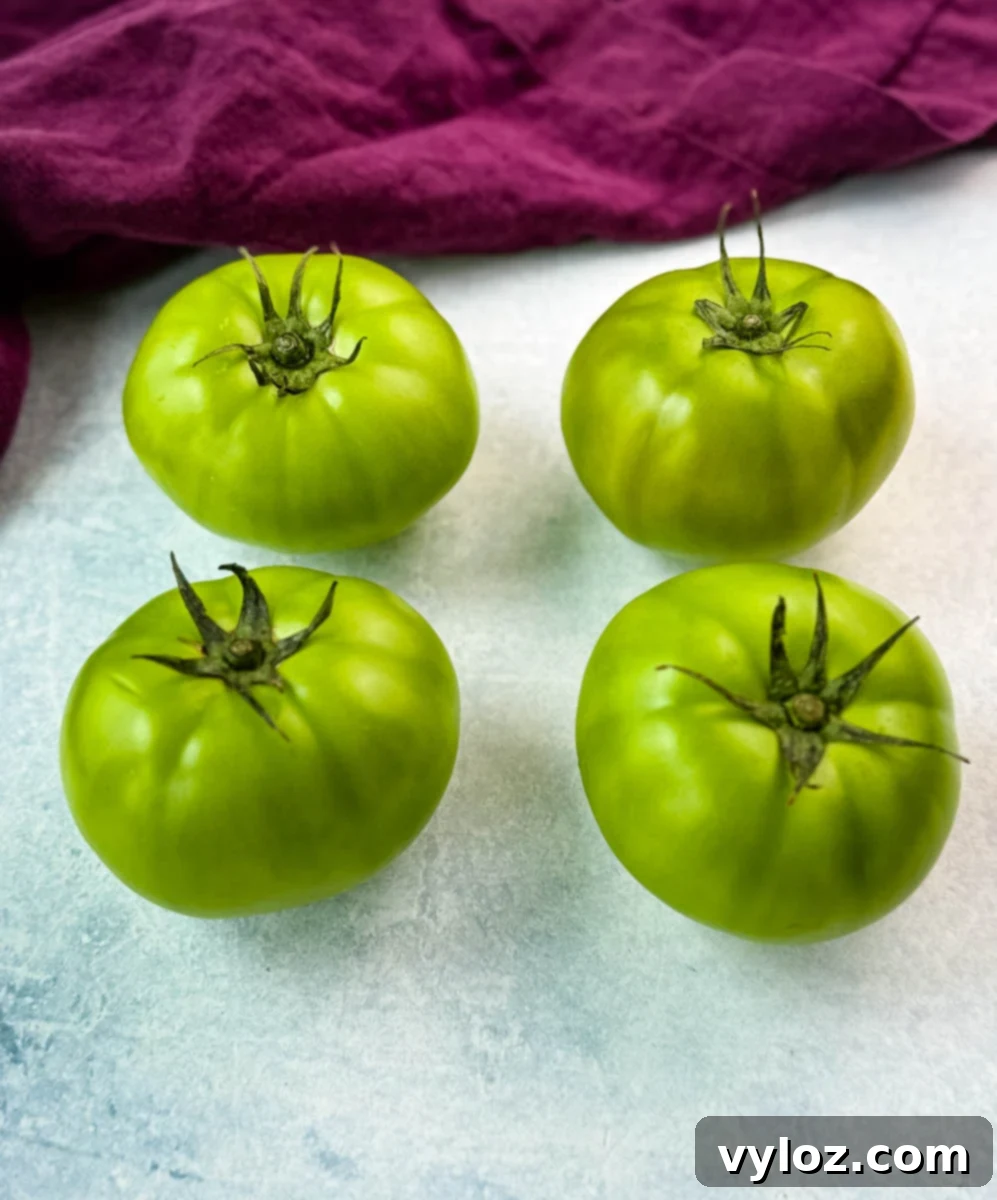 Southern Charm in Every Slice 3 Four whole, unripe green tomatoes with stems intact placed on a white surface next to a purple kitchen towel—perfect for frying.