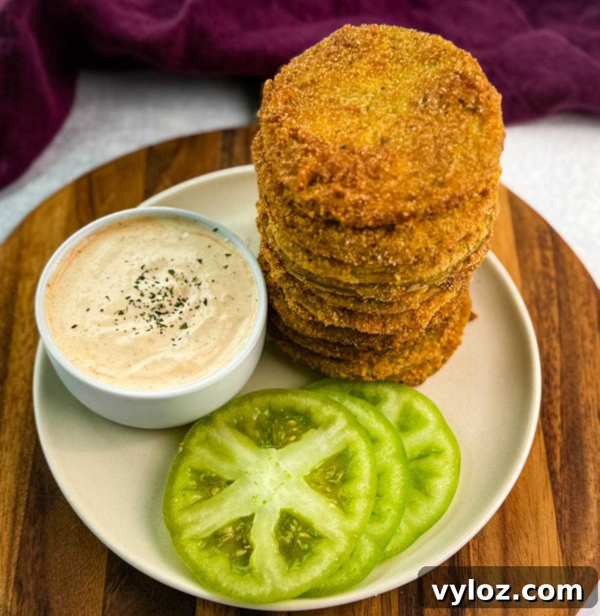 Southern Charm in Every Slice 9 Tall stack of fried green tomatoes served on a white plate with creamy dipping sauce and fresh tomato slices, all on a wooden board with a purple kitchen towel nearby.