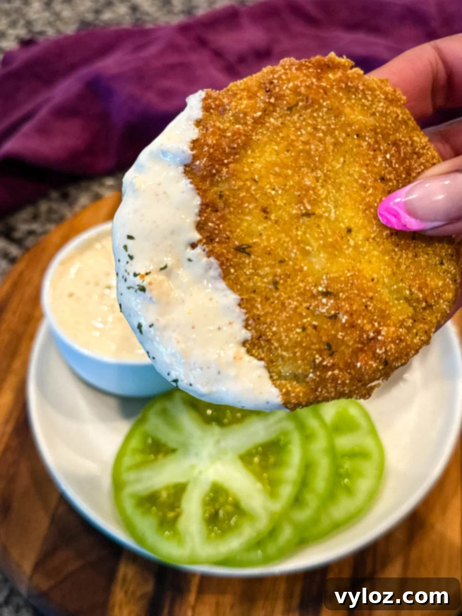 Easy Homemade Remoulade 5 Close-up of a hand holding a crispy, golden-brown fried green tomato partially dipped in creamy white sauce, with sliced raw green tomatoes and dipping sauce in the background.