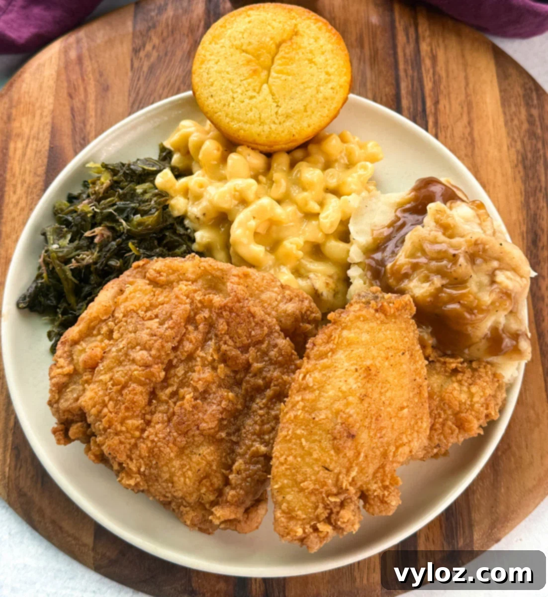 Overhead view of a full Southern dinner plate with golden fried chicken, creamy mac and cheese, collard greens, mashed potatoes with brown gravy, and a round cornbread muffin.