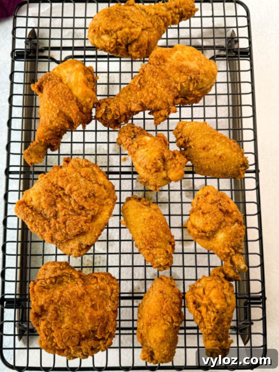 Multiple pieces of fried chicken spread across a wire rack for draining, showing their crispy, golden crust. Includes thighs, drumsticks, and wings.
