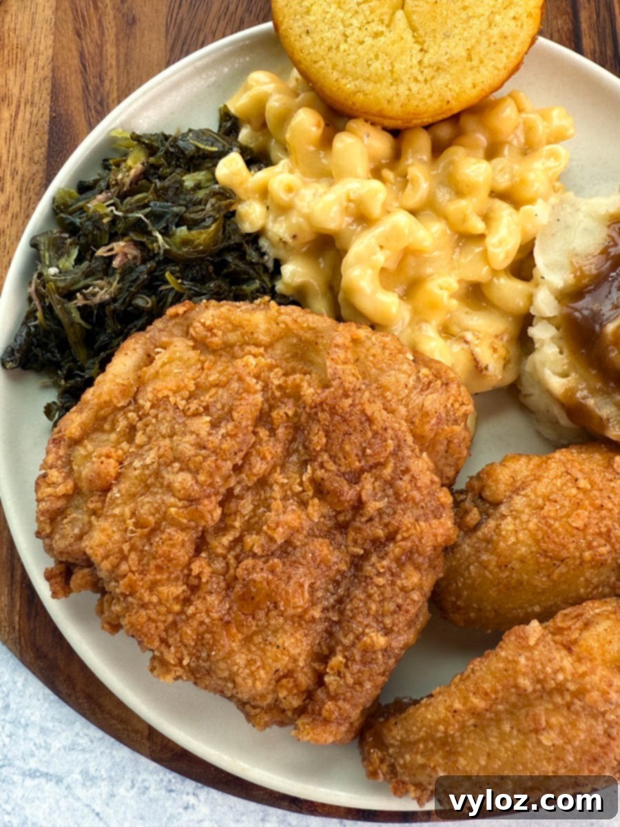 A plate of Southern comfort food featuring a large fried chicken thigh, collard greens, creamy mac and cheese, mashed potatoes with gravy, and a golden cornbread muffin.