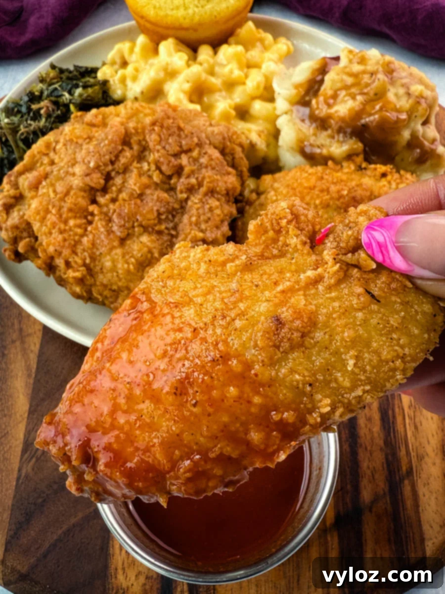 Close-up of a hand holding a freshly fried chicken wing dipped in hot sauce, with a plate in the background featuring fried chicken, mac and cheese, collard greens, mashed potatoes and gravy, and a cornbread muffin.