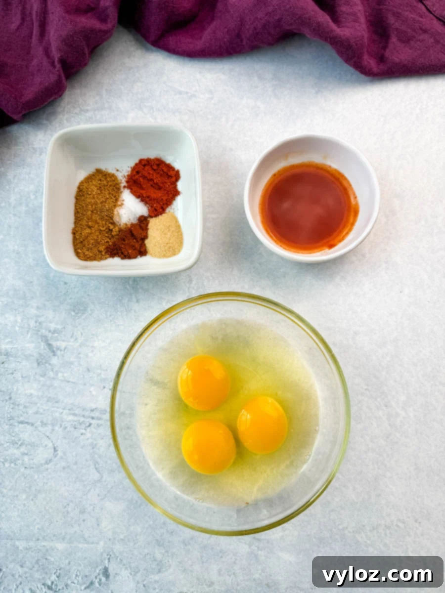 Three raw eggs in a glass bowl, with a dish of Creole seasoning, paprika, garlic powder, and cayenne pepper next to a small bowl of hot sauce—ingredients prepped for the egg wash and seasoning.