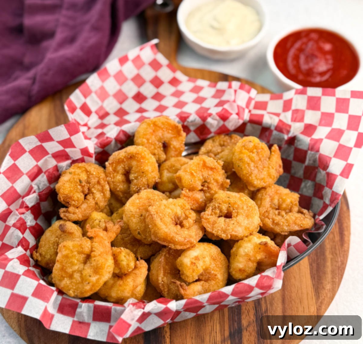 Front-focused shot of crispy fried shrimp piled high in a checkered basket, with sauce cups slightly out of focus in the background.