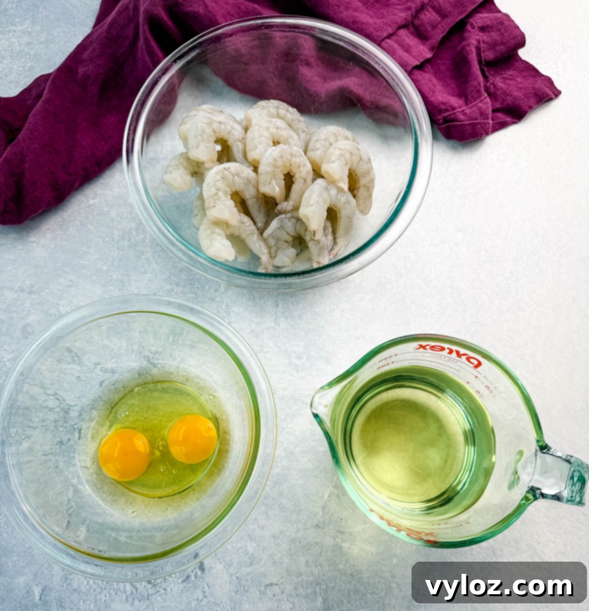Overhead shot of prep ingredients for fried shrimp including raw peeled shrimp, cracked eggs in a bowl, and oil in a measuring cup.