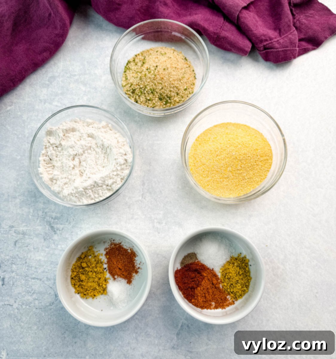 Overhead view of six small glass bowls showing individual ingredients for breading: flour, cornmeal, breadcrumbs, and seasonings, arranged on a light background with a purple towel.