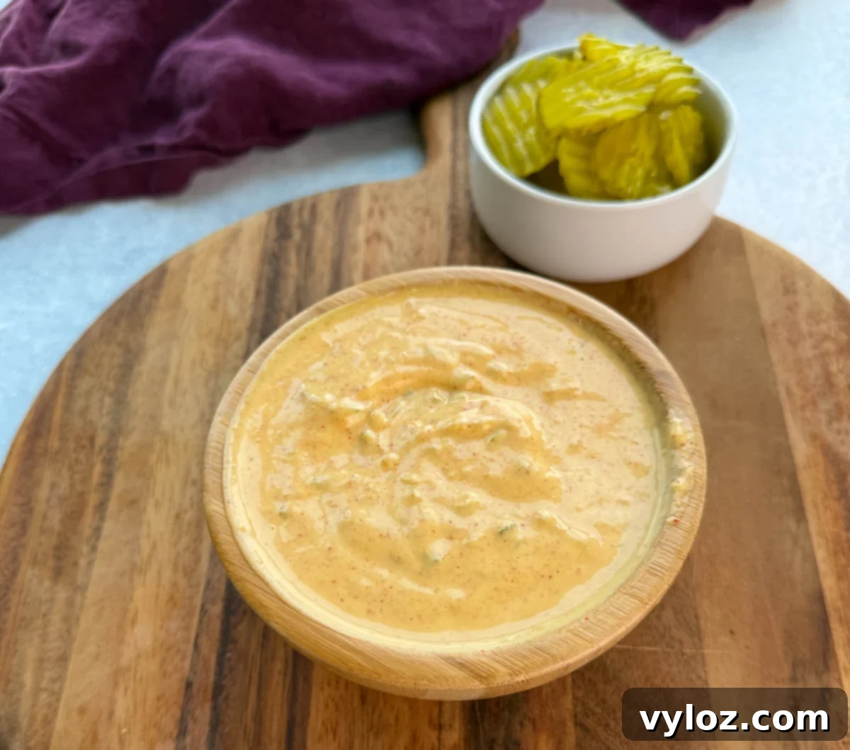 A wooden bowl filled with creamy Big Mac sauce on a wooden board, with a small white bowl of sliced pickles in the background.