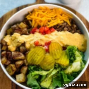 Top-down view of a Big Mac bowl with roasted potatoes, ground beef, shredded cheddar, chopped tomatoes, lettuce, pickles, and a drizzle of homemade sauce, served on a wooden board.