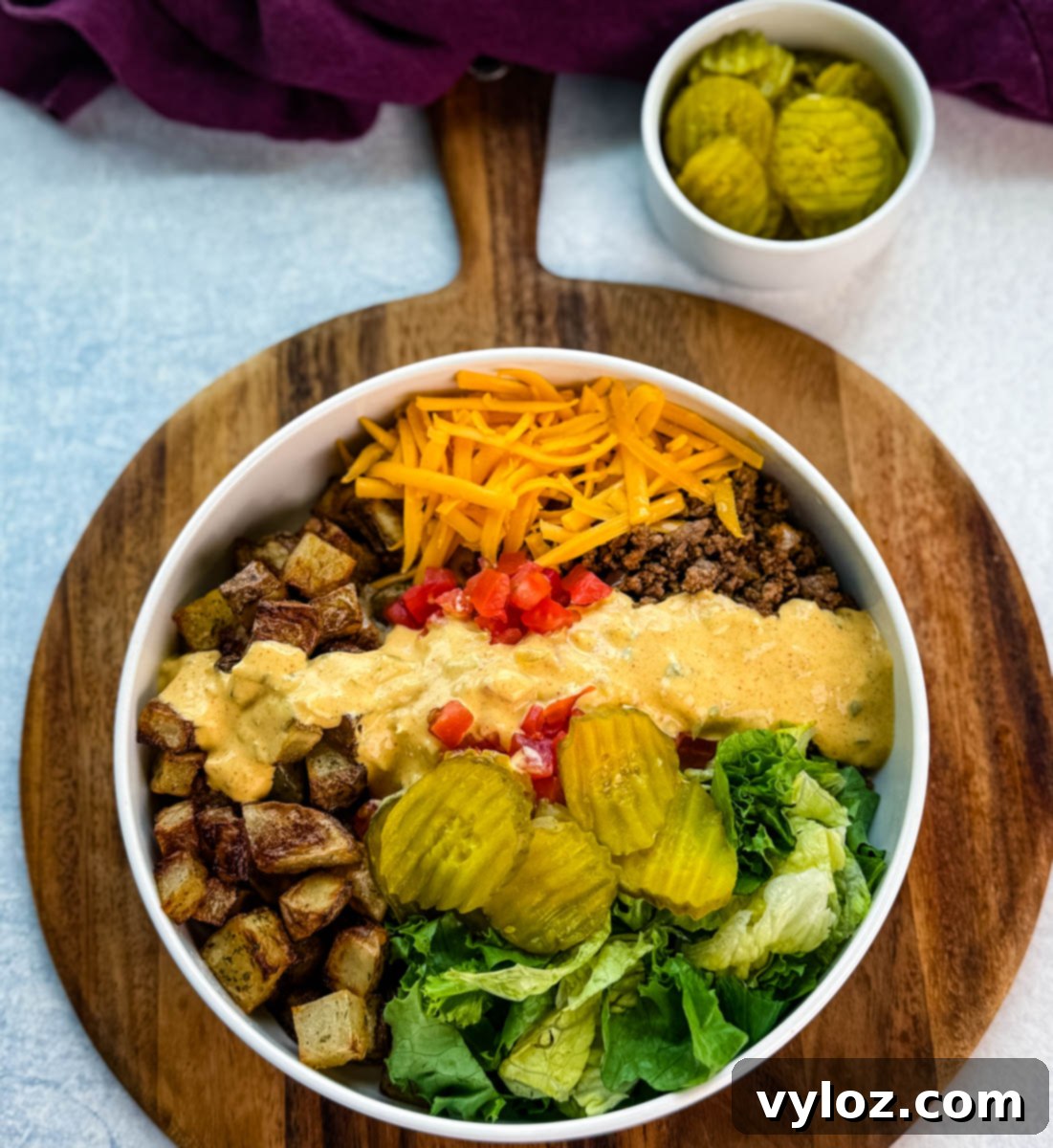 Wide overhead image of a fully assembled Big Mac bowl on a wooden serving board, with a small dish of sliced pickles in the background.