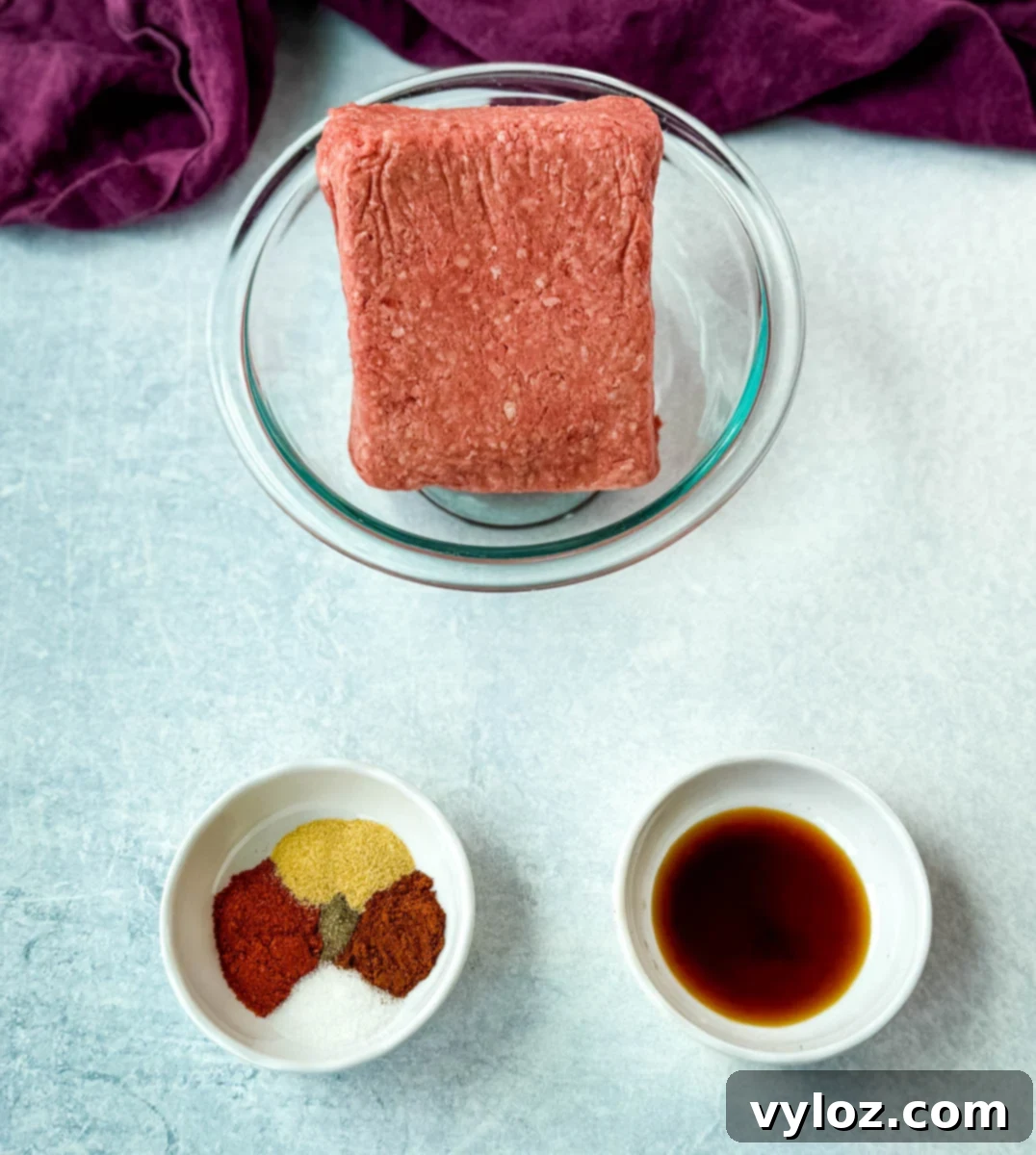 A block of raw ground beef in a glass bowl, with two small bowls of seasoning mix and Worcestershire sauce arranged nearby on a light countertop.