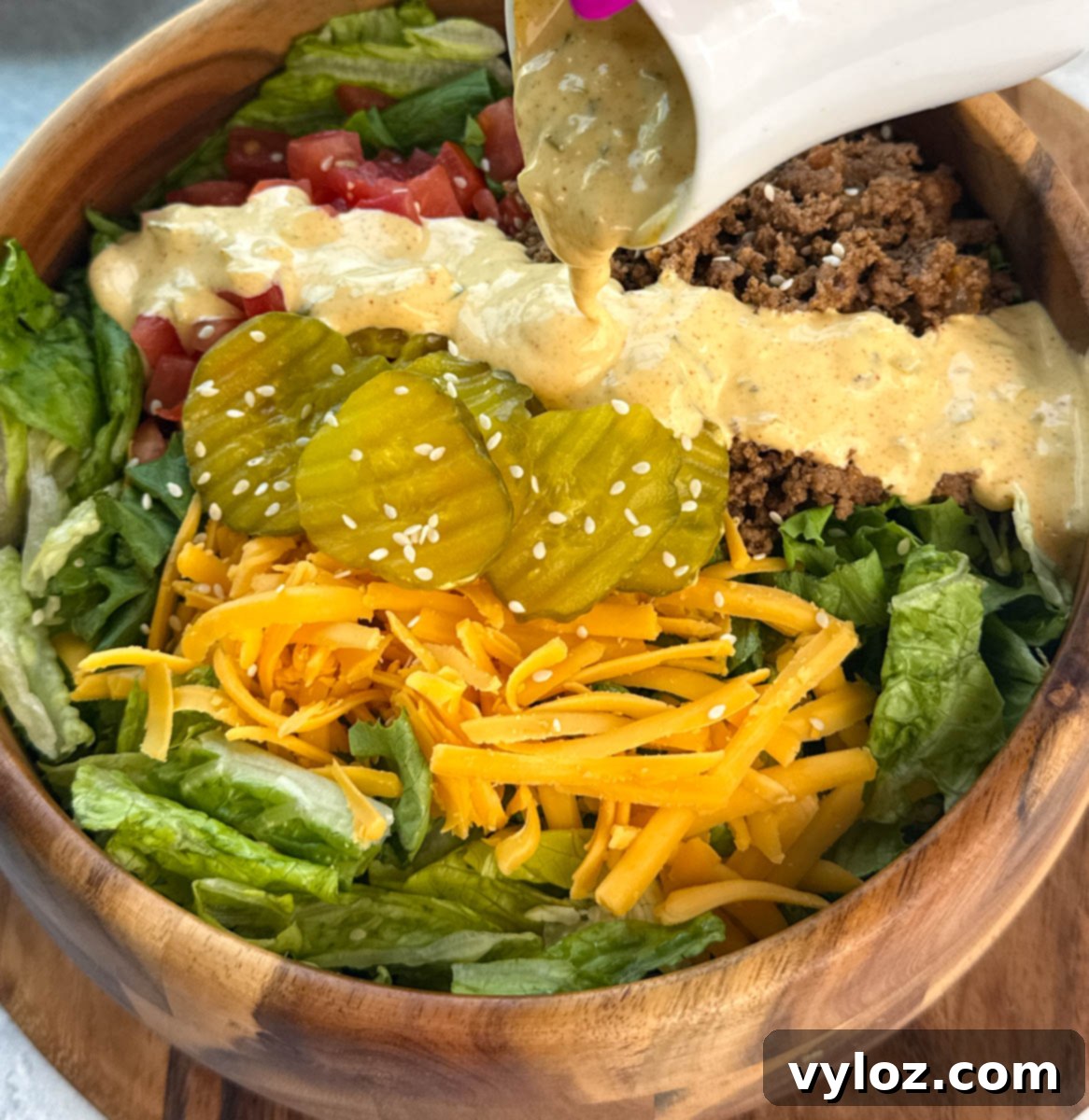 Big Mac salad in a wooden bowl with sauce being poured on top from a small white pitcher, highlighting fresh pickles, cheddar, lettuce, and tomatoes.