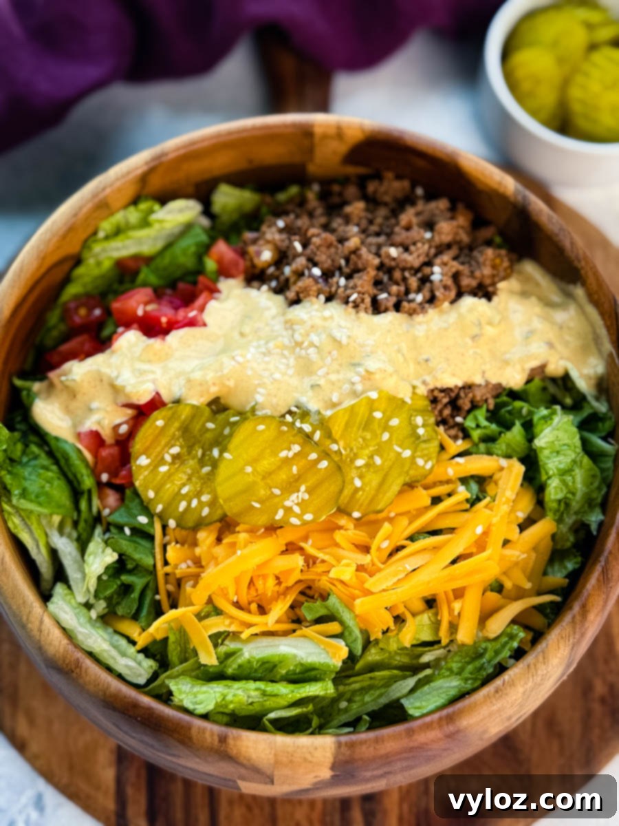 Top-down view of a colorful Big Mac salad in a wooden bowl, featuring layers of ground beef, shredded lettuce, cheddar cheese, pickles, tomatoes, and sauce with sesame seeds.