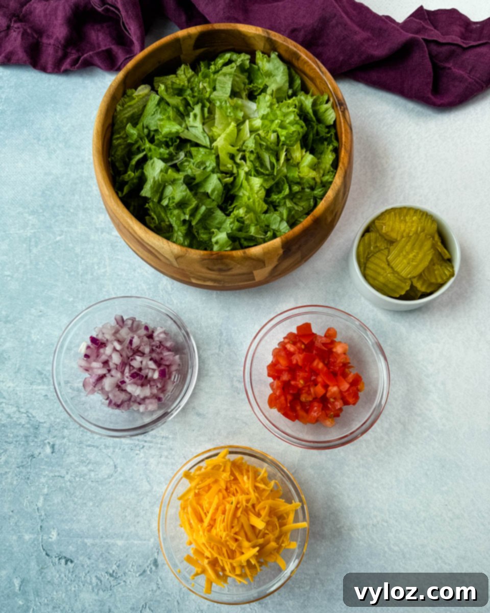 Ingredient prep for Big Mac bowls, featuring shredded lettuce in a wooden bowl, and smaller glass bowls of diced red onion, tomatoes, shredded cheddar cheese, and sliced pickles.