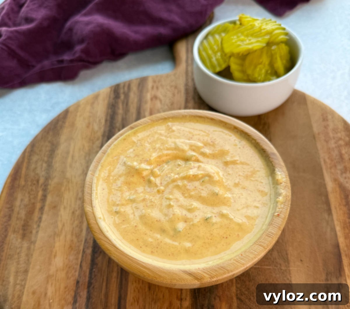 A wooden bowl filled with creamy Big Mac sauce on a wooden board, with a small white bowl of sliced pickles in the background.
