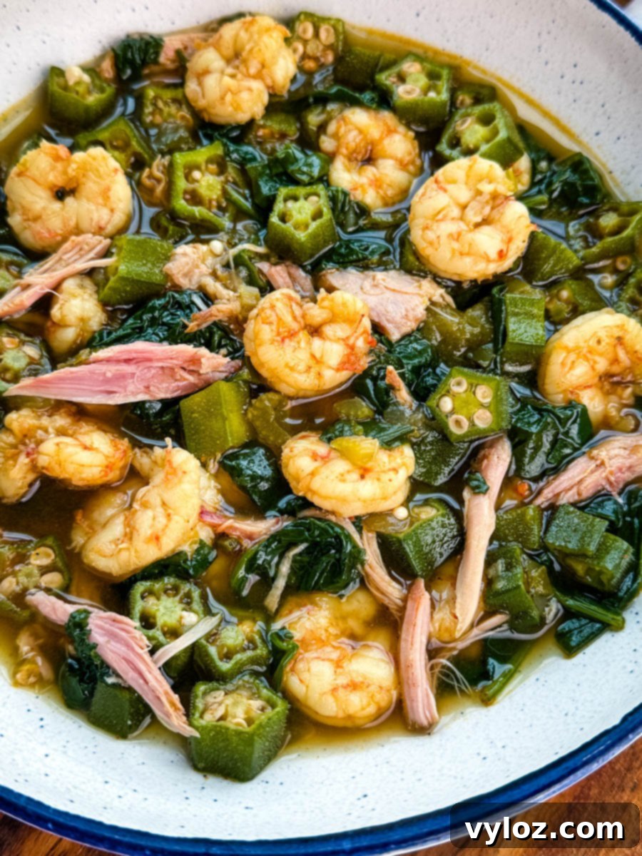 Close-up of okra soup showing shrimp, tender okra, shredded smoked turkey, and wilted greens in a rich broth.