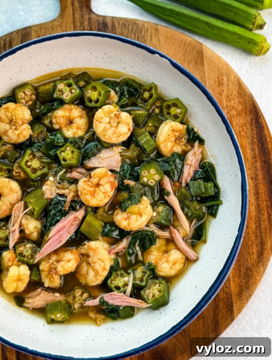 Bowl of okra soup with shrimp, smoked turkey pieces, and collard greens in broth, served on a wooden board with fresh okra nearby.