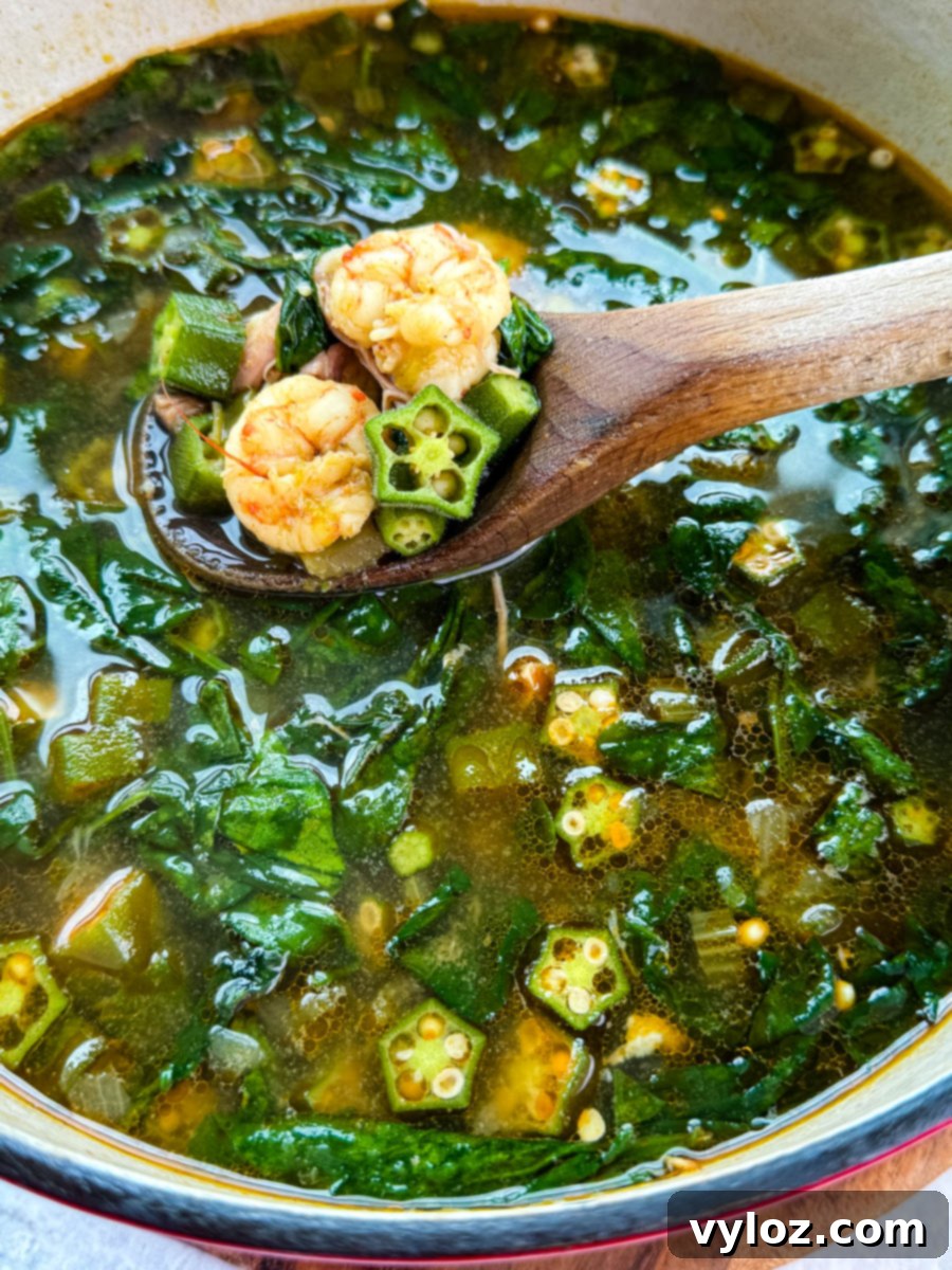 Wooden spoon lifting shrimp, okra, and spinach from a simmering pot of okra soup in seasoned broth.