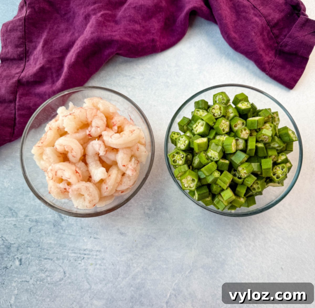 Two glass bowls side by side—one filled with raw peeled shrimp, the other with freshly chopped okra. A purple kitchen towel is in the background.
