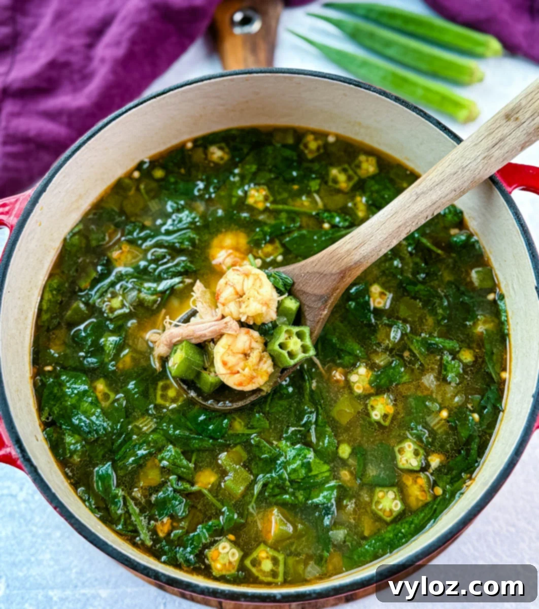 Wooden spoon lifting shrimp, okra, and spinach from a simmering pot of okra soup in seasoned broth.