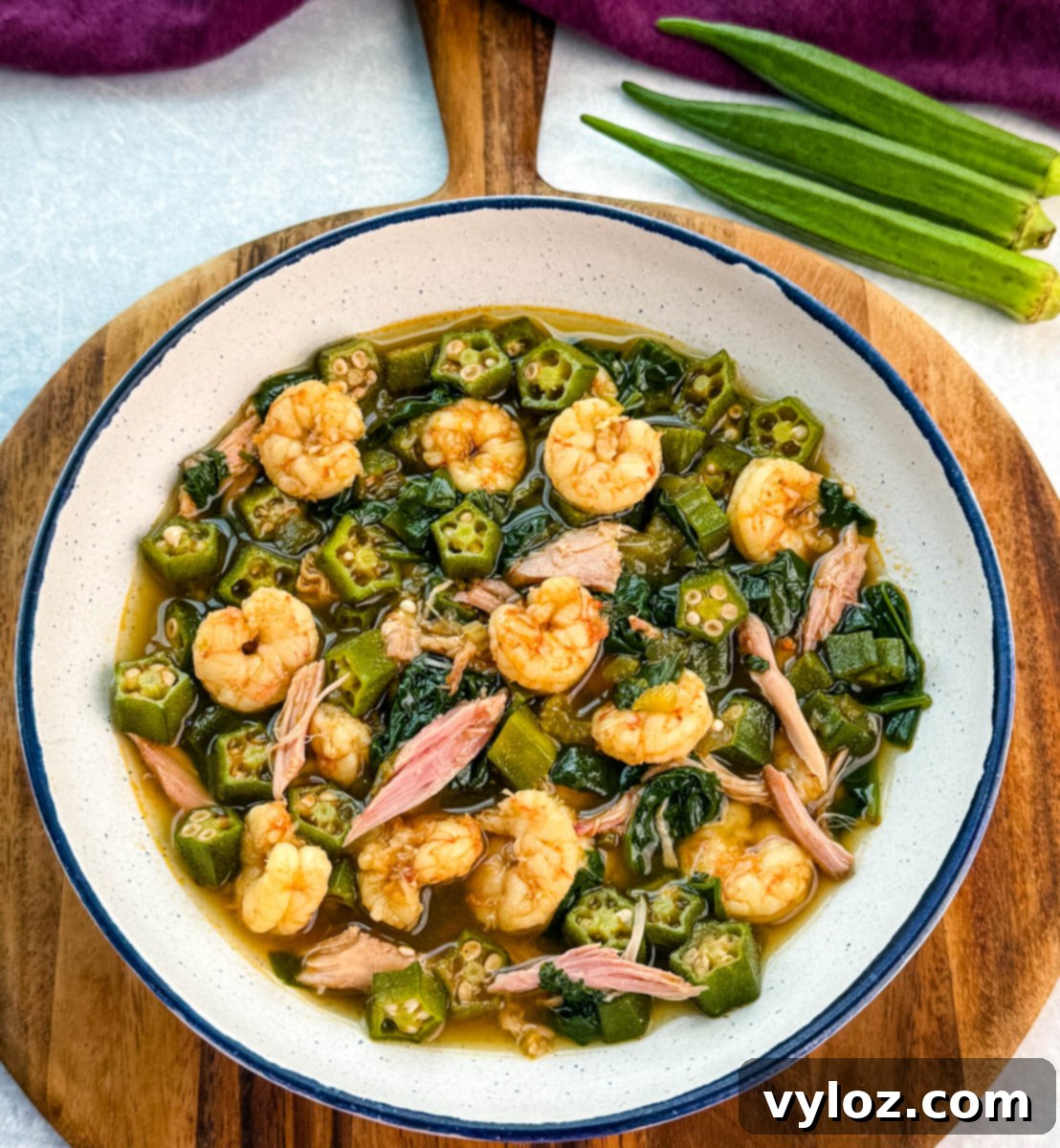 Overhead view of a bowl of okra soup filled with shrimp, okra, and shredded smoked turkey, placed on a wooden board with whole okra to the side.
