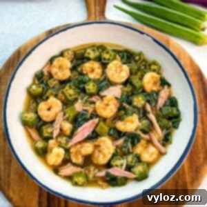 Bowl of okra soup filled with shrimp, chunks of smoked turkey, spinach, and okra in a rich broth, served on a wooden board with fresh okra in the background.