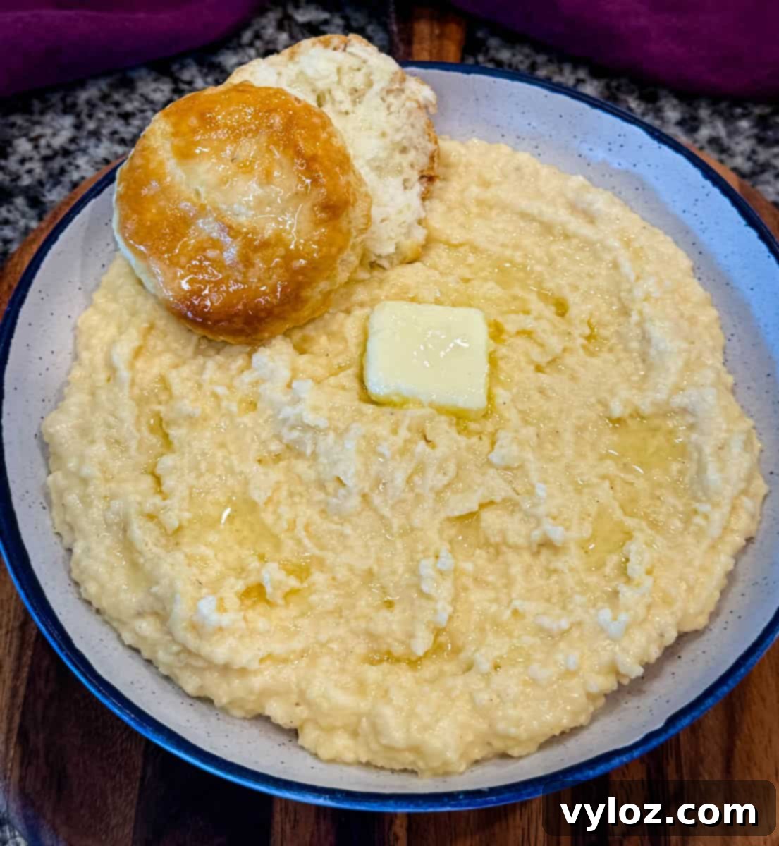 Close-up of creamy stone ground cheese grits in a speckled bowl, topped with a pat of melting butter and a flaky biscuit on the side.
