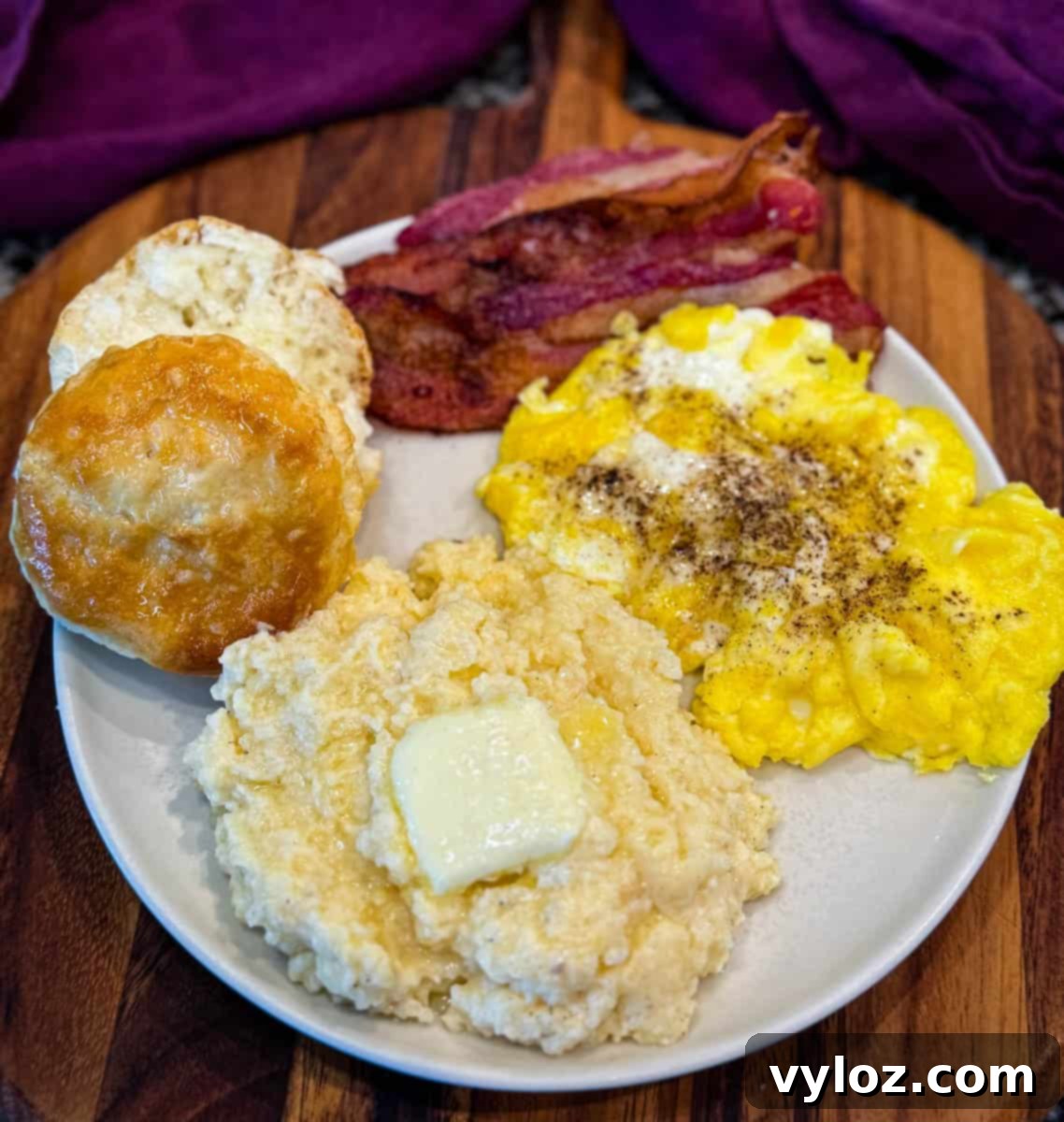 Southern-style breakfast plate with creamy cheese grits, scrambled eggs, crispy bacon, and a flaky biscuit, served on a white plate with a wooden background.