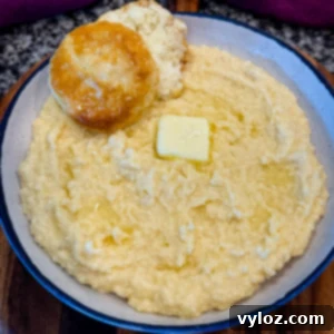 Close-up of creamy stone ground cheese grits in a speckled bowl, topped with a pat of melting butter and a flaky biscuit on the side.