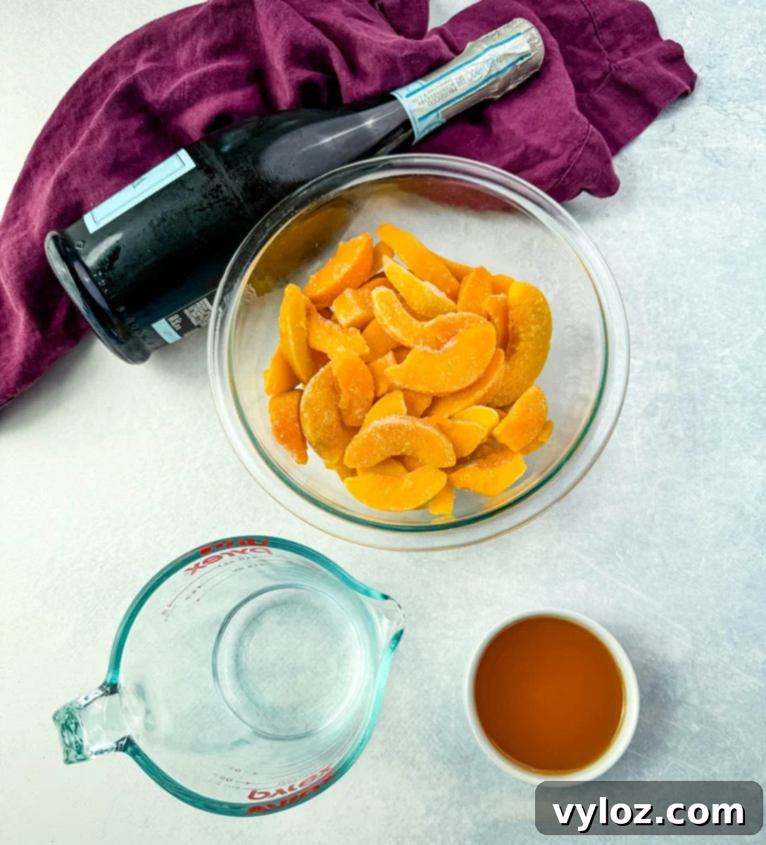 A selection of high-quality ingredients for a delightful peach Bellini slush: a bowl of vibrant frozen peach slices, a chilled bottle of Prosecco, a container of peach nectar, and fresh water, all arranged neatly on a kitchen countertop.