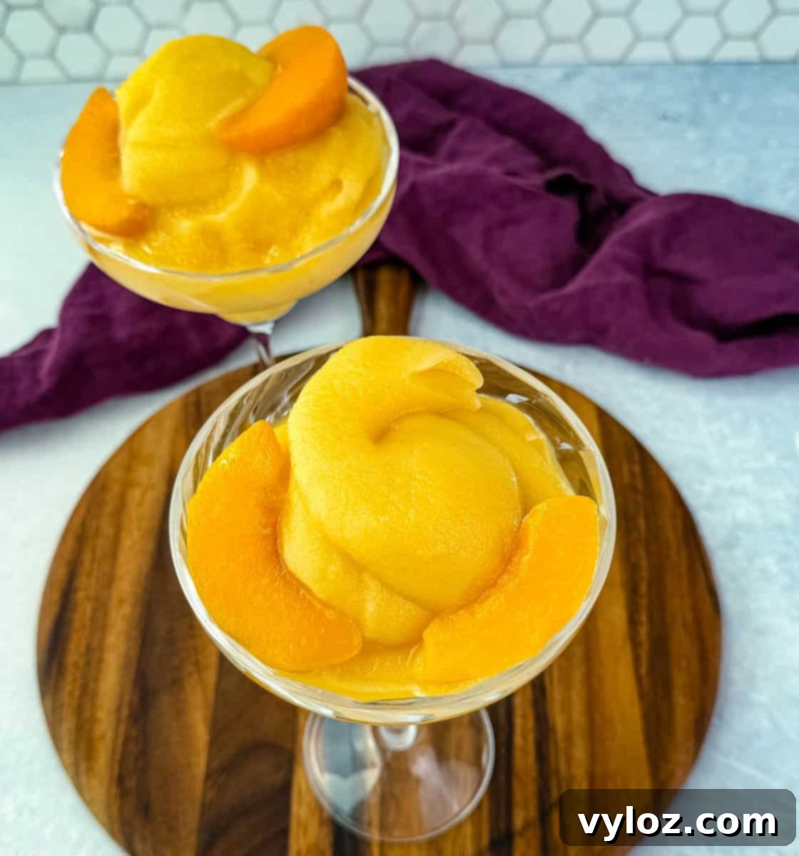Two cocktail glasses featuring the thick, frosted peach Bellini slush, artfully garnished with fresh peach slices, elegantly presented on a rustic wooden board.