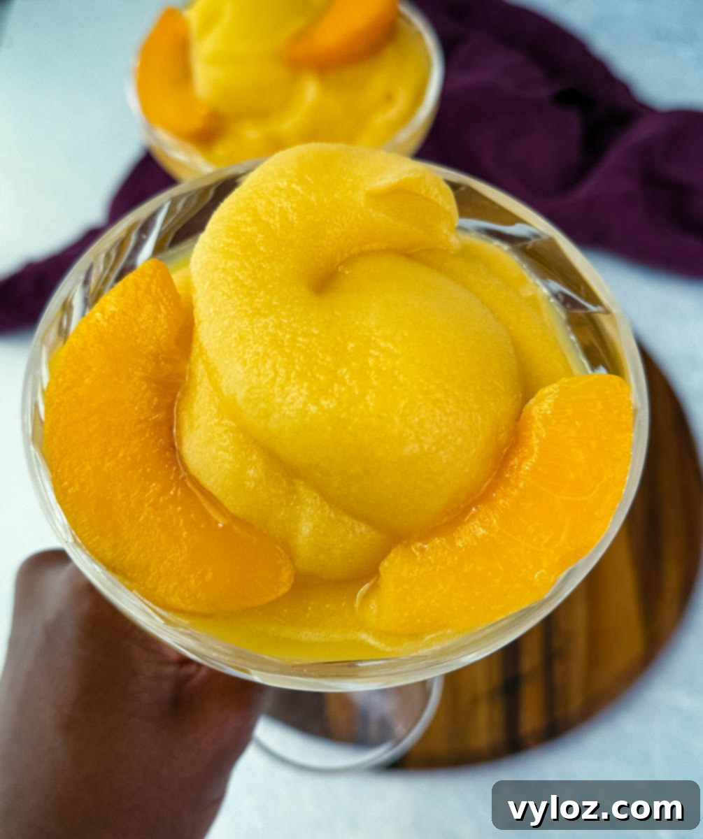 Close-up of a hand delicately holding a crystal glass, showcasing the creamy, vibrant frozen peach Bellini slush, perfectly garnished with a thin, fresh peach slice.