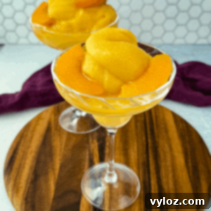 Two cocktail glasses filled with thick peach Bellini slush, garnished with peach slices, placed on a wooden board.