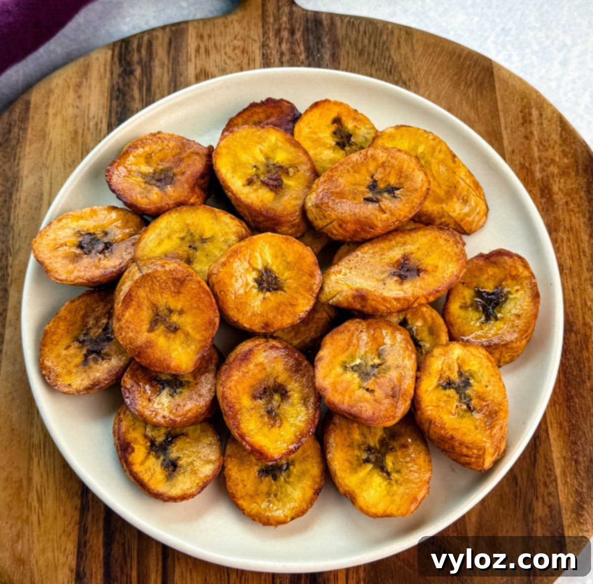 Plate of golden, crispy fried plantains stacked on a cream-colored plate set over a wooden board.