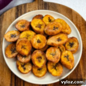 Plate of golden, crispy fried plantains stacked on a cream-colored plate set over a wooden board.