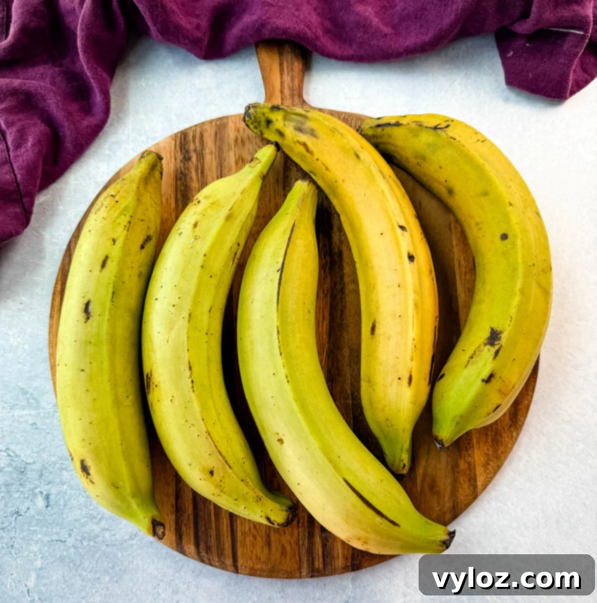 Partially ripe plantains with green and yellow skin on a round wooden board, ready for slicing.