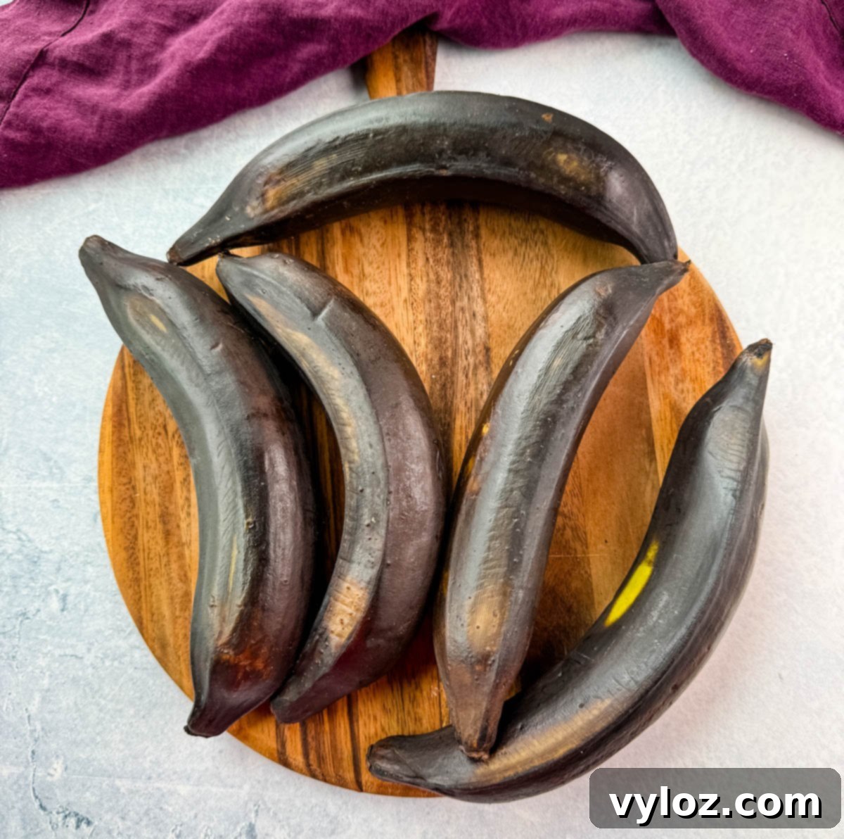 Fully ripened, dark black plantains on a round wooden board with a purple cloth.