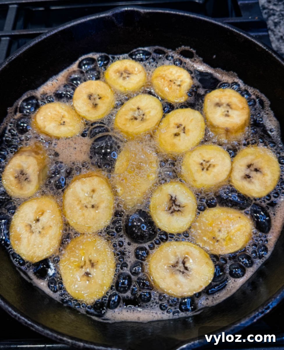 Raw plantain slices bubbling in oil in a black skillet, showing the start of the frying process.