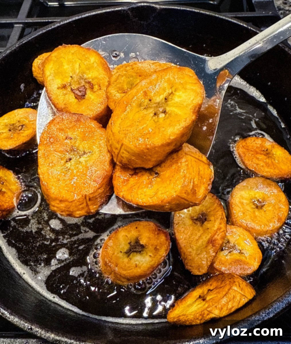 Sliced sweet plantains frying in hot oil in a black cast iron skillet, with golden edges just beginning to crisp.