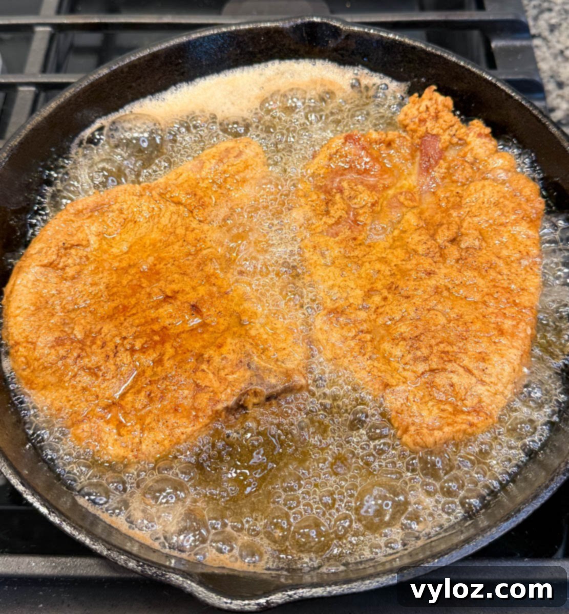 Crispy Southern Fried Pork Chops 10 Two bone-in pork chops frying in hot oil in a cast iron skillet, with visible bubbles around the edges, indicating proper frying temperature.