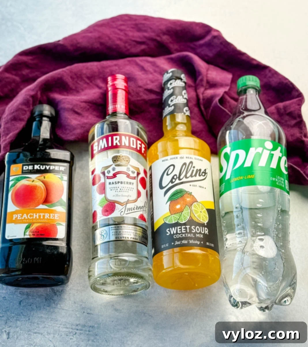 Boozy Bear Shots 3 Four drink ingredients laid side by side on a light surface with a purple cloth in the background: DeKuyper Peachtree peach schnapps, Smirnoff raspberry vodka, Collins sweet and sour cocktail mix, and a bottle of Sprite lemon-lime soda.