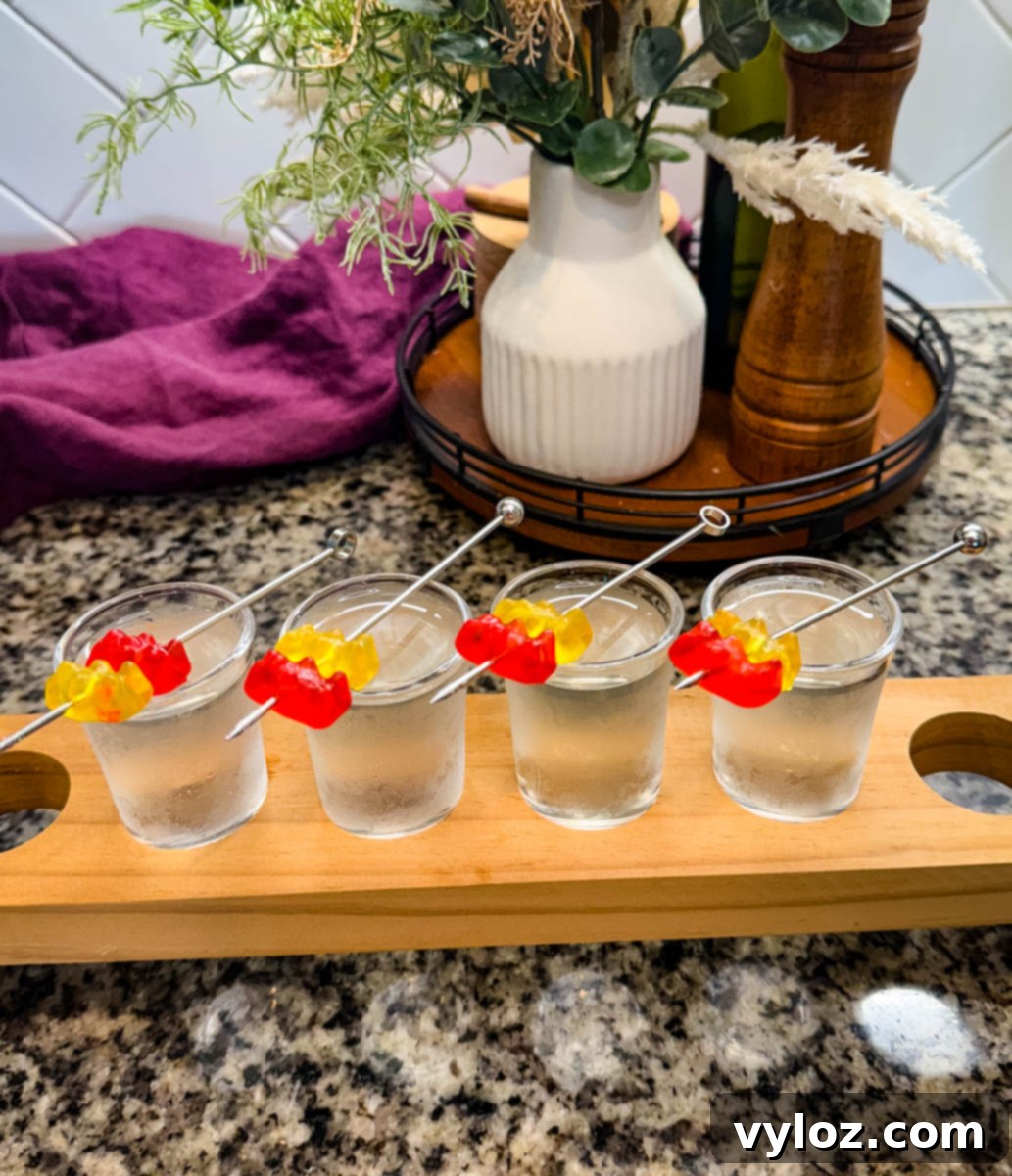 Boozy Bear Shots 6 Gummy bear cocktail shots placed on a wooden flight board in a kitchen setting. Each shot is garnished with red and yellow gummy bears, and kitchen decor with greenery is visible in the background.