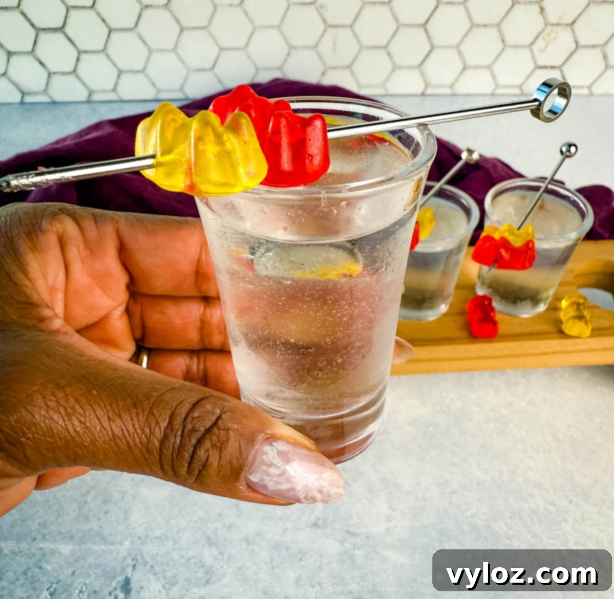 Boozy Bear Shots 7 A hand holding a clear gummy bear cocktail shot garnished with red and yellow gummy bears on a metal pick. More shots and a purple napkin are visible in the background.