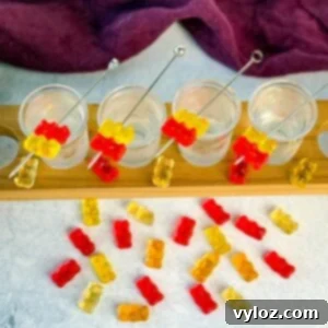 Boozy Bear Shots 8 Overhead view of four gummy bear cocktail shots in a wooden flight board, surrounded by scattered red, yellow, and orange gummy bears on a light surface.